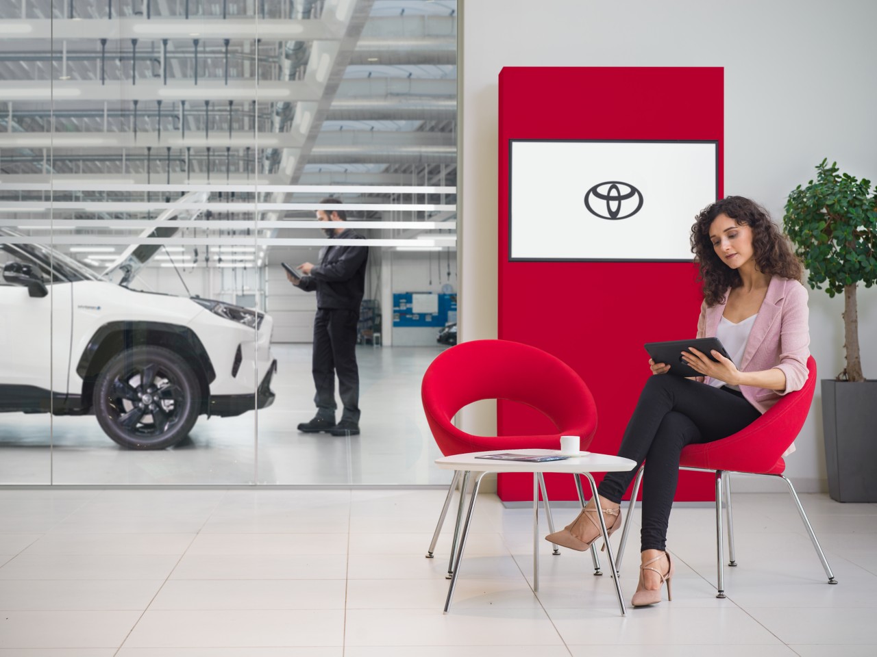 Žena sedí v zákaznickém prostoru Toyota Service. Zatímco sleduje tablet, na televizní obrazovce na pozadí je vidět logo Toyota.