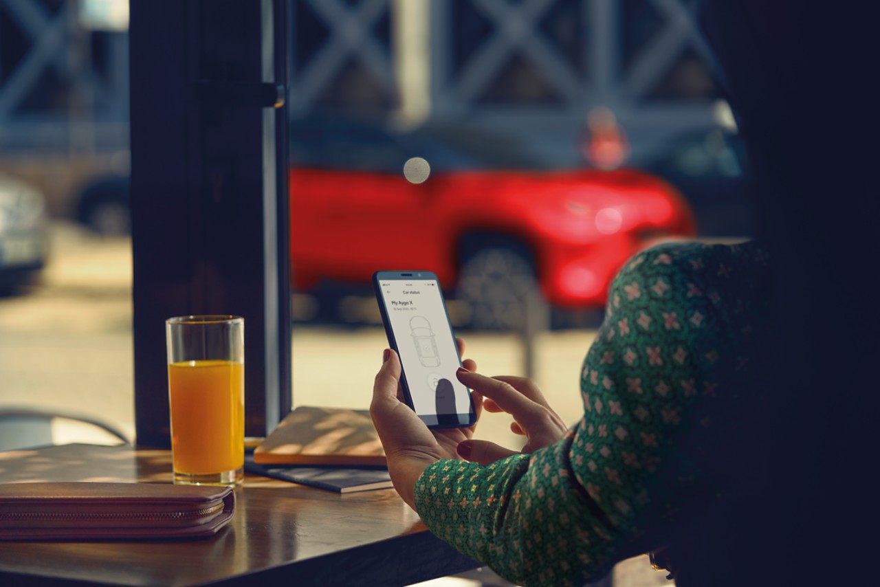 Žena sedí v kavárně. Zatímco používá aplikaci MyToyota na telefonu, na pozadí stojí zaparkována její Toyota Yaris Cross.